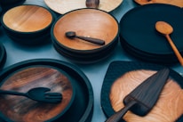 An assortment of wooden kitchenware including plates, bowls, spoons, ladles, and spatulas are arranged on a flat surface. The items are made from different shades of wood, with some pieces having a polished finish while others have a more rustic appearance.