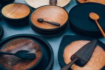 An assortment of wooden kitchenware including plates, bowls, spoons, ladles, and spatulas are arranged on a flat surface. The items are made from different shades of wood, with some pieces having a polished finish while others have a more rustic appearance.