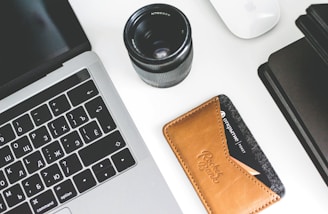 A workspace setup featuring a laptop with Cyrillic keyboard layout, a camera lens next to it, a leather card holder with a visible credit card, a white mouse, and a black notebook. The items are arranged neatly on a white surface.
