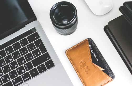 A workspace setup featuring a laptop with Cyrillic keyboard layout, a camera lens next to it, a leather card holder with a visible credit card, a white mouse, and a black notebook. The items are arranged neatly on a white surface.