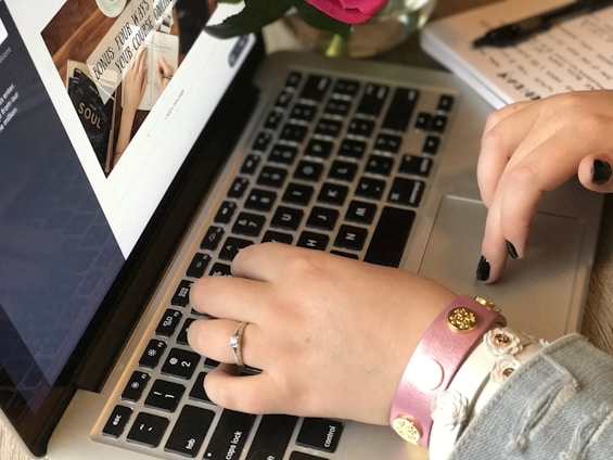A pair of hands typing on a laptop keyboard, one hand wearing several bracelets and a ring with black-painted nails. To the left, a webpage is open on the laptop screen displaying text and a book titled 'SOUL'. Nearby, a part of a notebook and a pen are visible, along with a flower in the background.