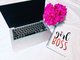 A laptop is open on a bed next to a notebook with the words 'girl boss' written on the cover in a stylish font. Beside them, there's a vase filled with vibrant pink flowers.