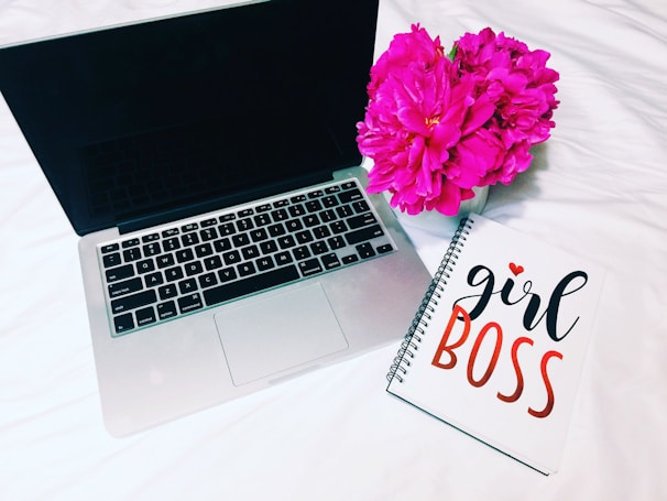 A laptop is open on a bed next to a notebook with the words 'girl boss' written on the cover in a stylish font. Beside them, there's a vase filled with vibrant pink flowers.