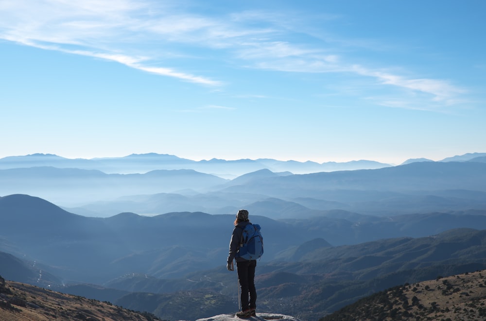 person wearing backpack standing on cliff in front of mountains during daytime