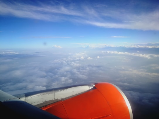 An airplane engine is visible in the foreground, with a bright orange color contrasting against a vast expanse of clouds. The sky above is a brilliant blue, dotted with patches of white clouds, creating a seamless horizon.