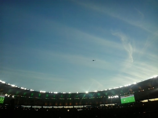 A large stadium is depicted with bright lights illuminating the stands, which are in the lower part of the image. There is a big screen displaying a sports event on the right side of the stadium. The sky above is vast and clear with a few wispy clouds and a helicopter is flying in the distance.