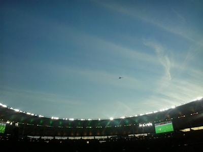 A large stadium is depicted with bright lights illuminating the stands, which are in the lower part of the image. There is a big screen displaying a sports event on the right side of the stadium. The sky above is vast and clear with a few wispy clouds and a helicopter is flying in the distance.