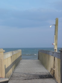 A wooden boardwalk leads directly to the ocean, with calm blue waves visible in the background. On the right side of the boardwalk, there is a metal shower fixture attached to a tall wooden pole. The sky above is mostly clear with a few clouds.
