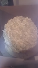 A round cake with an elaborate decoration of white frosting swirls, resembling flowers or rosettes. Small, pearl-like decorations are scattered across the frosting. The cake sits in what seems to be a cardboard box with a partially visible inscription at the bottom.