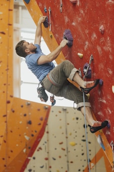 A person is actively rock climbing indoors on a vibrant climbing wall with various colorful handholds. The climber is focused and geared up with safety harness and rope, wearing a blue shirt and khaki shorts.