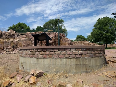 A stone well structure surrounded by rocky terrain and dry grasses is situated in an outdoor setting with trees and a clear sky with some clouds. A metal structure resembling a lamp post and information stand is mounted on the well.