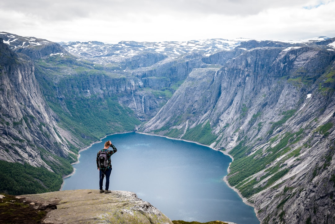 man in black jacket with black backpack standing on top of cliff above a lake at daytime