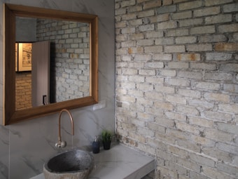 A bathroom sink area featuring a stone basin and a copper faucet is set against a marble countertop. A large wooden-framed mirror hangs above the sink, reflecting a partially open door and framed art on an exposed brick wall. The walls are a mix of marble and brick textures, with a small potted plant and soap dispenser on the counter.