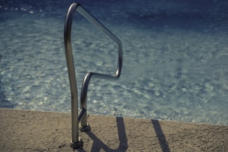 A metallic pool ladder is positioned at the edge of a swimming pool, casting a shadow on the textured poolside surface. The water in the pool is clear, reflecting sunlight with a slight ripple effect.