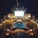 A luxury cruise ship deck featuring a pool surrounded by lounge chairs and warm ambient lighting. A large screen overlooks the area, showing an image against a dark night sky.