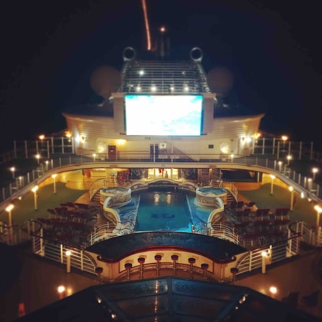 A luxury cruise ship deck featuring a pool surrounded by lounge chairs and warm ambient lighting. A large screen overlooks the area, showing an image against a dark night sky.