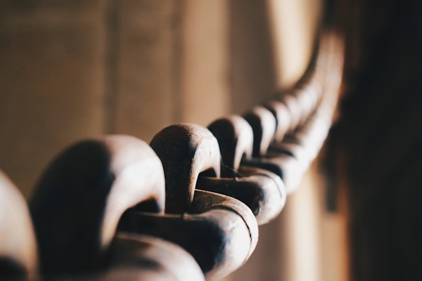 A close-up view of a metal chain links with a shallow depth of field, creating a blurred background. The chain appears to be well-worn, with a slightly rusty texture, and the light casts a warm glow over the metal surface.