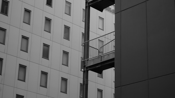 A grayscale architectural scene depicting two modern buildings, one with a grid-like pattern of square windows and another with a solid facade. A metal structure, possibly a balcony or walkway, connects the buildings.