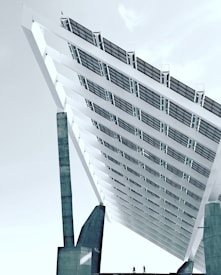 A large, angular solar panel structure is positioned against a cloudy sky. The architecture features massive supporting columns and panels that stretch upwards at a steep angle. Two small silhouettes of people walking or cycling can be seen at the base, emphasizing the scale of the structure.