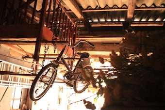 A vintage bicycle is hanging from the ceiling of a cluttered garage or workshop, secured with chains. There are wooden structures and metal elements around, including a ladder leaning against the wall. The space is dimly lit, creating an atmospheric and nostalgic feel.