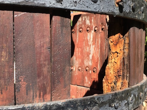 Close-up of a weathered wooden structure with metal banding and visible rust. The wood shows signs of age with peeling paint and an exposed, textured surface.