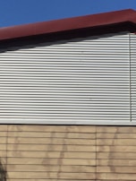 A building exterior features a combination of corrugated metal siding on the upper portion and horizontal wooden panels on the lower portion. The roof is angled and has a dark red trim. The sky is clear and blue, providing a contrasting backdrop.
