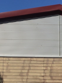 A building exterior features a combination of corrugated metal siding on the upper portion and horizontal wooden panels on the lower portion. The roof is angled and has a dark red trim. The sky is clear and blue, providing a contrasting backdrop.