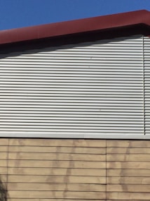 A building exterior features a combination of corrugated metal siding on the upper portion and horizontal wooden panels on the lower portion. The roof is angled and has a dark red trim. The sky is clear and blue, providing a contrasting backdrop.