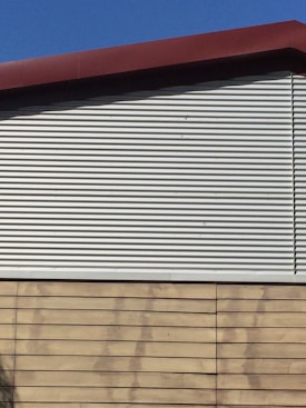 A building exterior features a combination of corrugated metal siding on the upper portion and horizontal wooden panels on the lower portion. The roof is angled and has a dark red trim. The sky is clear and blue, providing a contrasting backdrop.