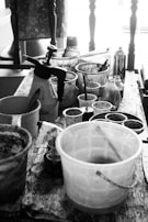 A black and white image of a wooden table cluttered with various art supplies including paint containers, brushes, spray bottles, and cups. The background features a chair with a cushioned seat and several vertical wooden spindles.