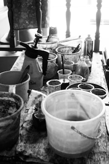 A black and white image of a wooden table cluttered with various art supplies including paint containers, brushes, spray bottles, and cups. The background features a chair with a cushioned seat and several vertical wooden spindles.