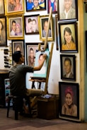 An artist sits on a stool, working on a portrait at an easel surrounded by numerous framed paintings on the wall. The images depict various people, some smiling and others with neutral expressions. The setting has a cozy, artistic vibe.