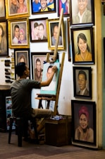 An artist sits on a stool, working on a portrait at an easel surrounded by numerous framed paintings on the wall. The images depict various people, some smiling and others with neutral expressions. The setting has a cozy, artistic vibe.