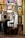 An artist sits on a stool, working on a portrait at an easel surrounded by numerous framed paintings on the wall. The images depict various people, some smiling and others with neutral expressions. The setting has a cozy, artistic vibe.