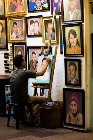 An artist sits on a stool, working on a portrait at an easel surrounded by numerous framed paintings on the wall. The images depict various people, some smiling and others with neutral expressions. The setting has a cozy, artistic vibe.