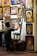 An artist sits on a stool, working on a portrait at an easel surrounded by numerous framed paintings on the wall. The images depict various people, some smiling and others with neutral expressions. The setting has a cozy, artistic vibe.