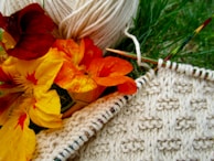 A close-up of a knitting project featuring cream-colored yarn and needles, alongside vibrant orange and yellow flowers. The knitted fabric displays a textured pattern, partially completed on the needles. Everything is placed on grass, creating a natural backdrop.