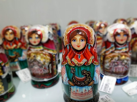 Colorful nesting dolls with intricate and ornate designs are displayed, each featuring a traditional painted woman in vibrant clothing with detailed patterns, including reds, greens, and golds. The dolls are arranged closely together, showcasing the craftsmanship and cultural artistry.