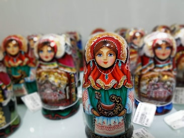 Colorful nesting dolls with intricate and ornate designs are displayed, each featuring a traditional painted woman in vibrant clothing with detailed patterns, including reds, greens, and golds. The dolls are arranged closely together, showcasing the craftsmanship and cultural artistry.