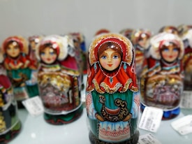 Colorful nesting dolls with intricate and ornate designs are displayed, each featuring a traditional painted woman in vibrant clothing with detailed patterns, including reds, greens, and golds. The dolls are arranged closely together, showcasing the craftsmanship and cultural artistry.