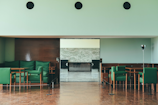 A modern interior space featuring green upholstered chairs arranged around wooden tables, with a wooden paneled wall and light mint-colored walls. The flooring is wooden, and there is a metal floor lamp to the right.