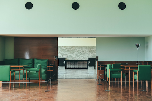 A modern interior space featuring green upholstered chairs arranged around wooden tables, with a wooden paneled wall and light mint-colored walls. The flooring is wooden, and there is a metal floor lamp to the right.