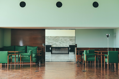 A modern interior space featuring green upholstered chairs arranged around wooden tables, with a wooden paneled wall and light mint-colored walls. The flooring is wooden, and there is a metal floor lamp to the right.