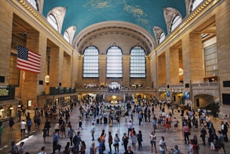 A bustling grand hall with a high arched ceiling painted with night sky imagery, large arched windows allowing natural light to filter in, and an American flag hanging prominently. The floor is occupied by numerous people walking or standing in groups. Chandeliers hang from the ceiling, adding a touch of elegance.