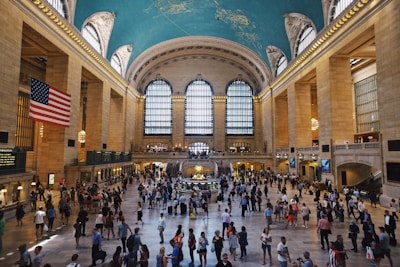 A bustling grand hall with a high arched ceiling painted with night sky imagery, large arched windows allowing natural light to filter in, and an American flag hanging prominently. The floor is occupied by numerous people walking or standing in groups. Chandeliers hang from the ceiling, adding a touch of elegance.