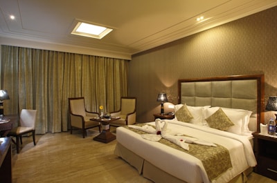 A well-decorated hotel room with warm lighting and neutral tones. The room features a large bed with decorative pillows and towel swans on it. There are two armchairs and a small table near the window with floor-length curtains. Nightstands on either side of the bed hold lamps with textured shades. A bottle of water and glasses are placed on one of the nightstands.