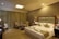 A well-decorated hotel room with warm lighting and neutral tones. The room features a large bed with decorative pillows and towel swans on it. There are two armchairs and a small table near the window with floor-length curtains. Nightstands on either side of the bed hold lamps with textured shades. A bottle of water and glasses are placed on one of the nightstands.