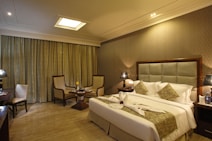 A well-decorated hotel room with warm lighting and neutral tones. The room features a large bed with decorative pillows and towel swans on it. There are two armchairs and a small table near the window with floor-length curtains. Nightstands on either side of the bed hold lamps with textured shades. A bottle of water and glasses are placed on one of the nightstands.