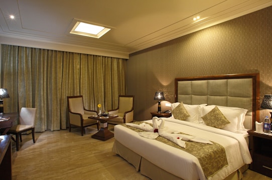 A well-decorated hotel room with warm lighting and neutral tones. The room features a large bed with decorative pillows and towel swans on it. There are two armchairs and a small table near the window with floor-length curtains. Nightstands on either side of the bed hold lamps with textured shades. A bottle of water and glasses are placed on one of the nightstands.