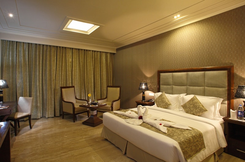 A well-decorated hotel room with warm lighting and neutral tones. The room features a large bed with decorative pillows and towel swans on it. There are two armchairs and a small table near the window with floor-length curtains. Nightstands on either side of the bed hold lamps with textured shades. A bottle of water and glasses are placed on one of the nightstands.
